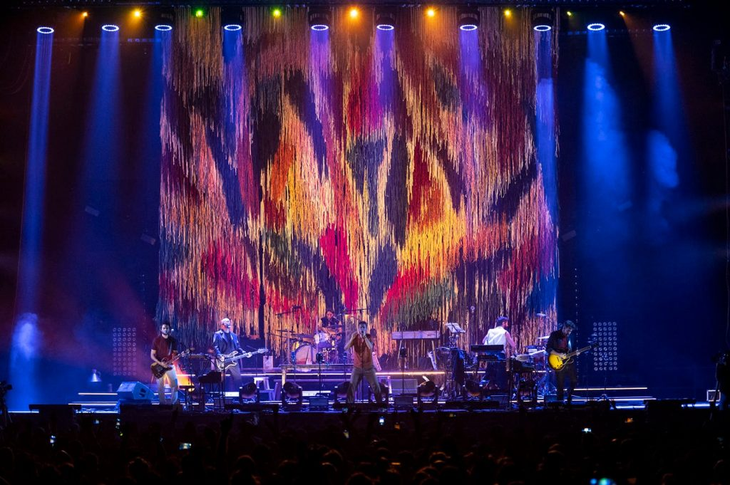 Imagen del escenario de un concierto de vetusta morla, donde aparece de fondo el telar encargado a graffica y que sería la imagen de la gira, con retales de colores